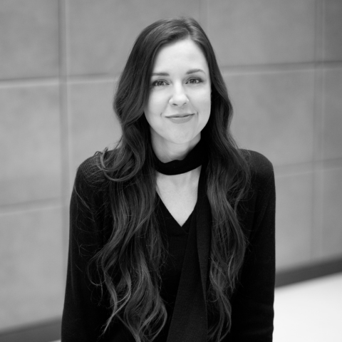 Portrait en noir et blanc de Carla Buzzasi, aux cheveux longs et foncés, portant une veste noire, devant un mur gris.