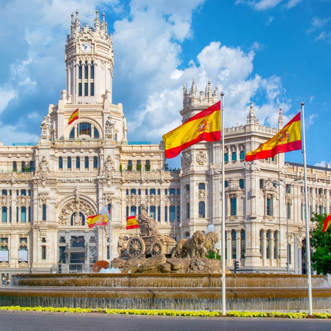 View of the Cibeles Fountain in Madrid with the Cibeles Palace in the background and several Spanish flags waving in the wind.