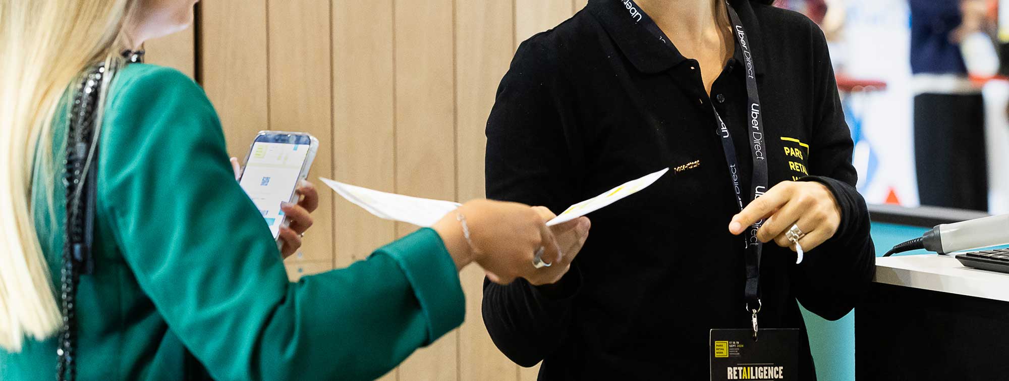 Échange entre deux personnes au stand d'accueil, l'une remettant un document et l'autre consultant un smartphone.