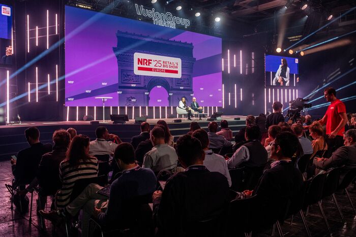 Photograph of a conference at Siec 2025, featuring an audience of business professionals seated in front of a purple-lit stage, with several speakers participating in a panel discussion and a large screen in the background.