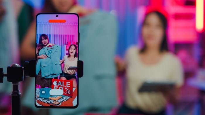 Retail Media illustration featuring a smartphone recording a content creator showcasing promotional products during a livestream shopping session, in a retail environment with vibrant pink and blue lighting.