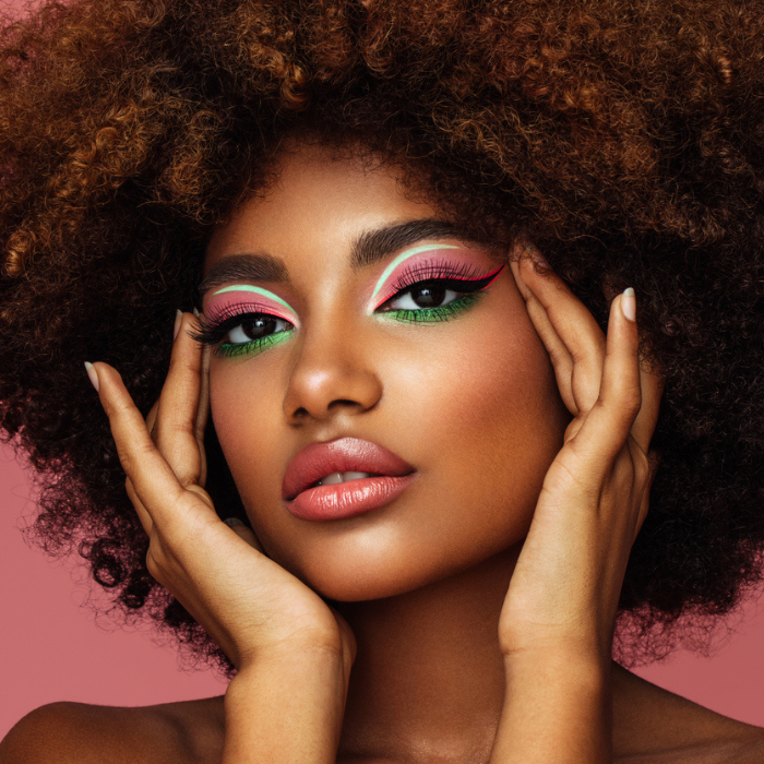 Beauty portrait of a woman with voluminous natural afro hair, wearing vibrant artistic makeup with turquoise and pink eyeshadow, gently framing her face with her hands against a pink background.