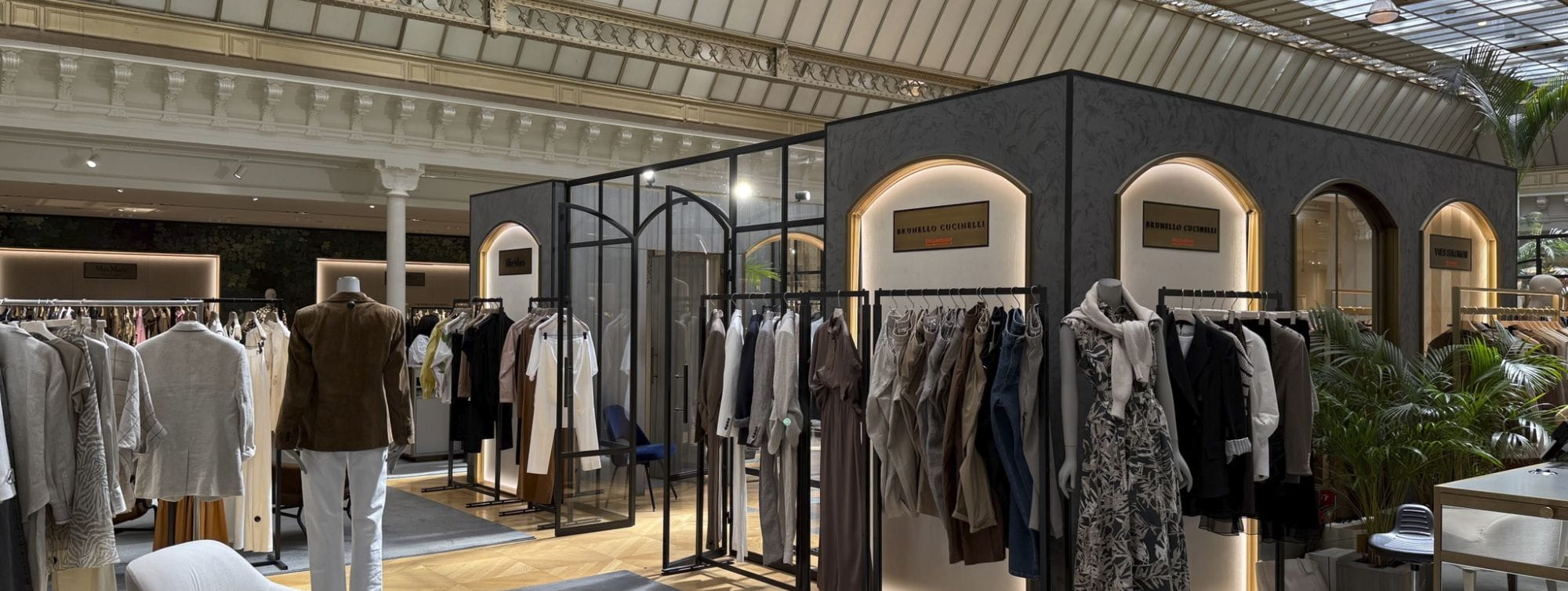 Vue d’un espace de vente haut de gamme avec des présentoirs de vêtements élégants, des cabines vitrées et des enseignes de marques de luxe comme Brunello Cucinelli et Yves Salomon, sous une verrière lumineuse au design architectural raffiné.