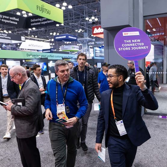 Visite guidée du salon NRF 2025, un intervenant tient un panneau "The New Connected Store Journey" en tête de groupe, au milieu des exposants.
