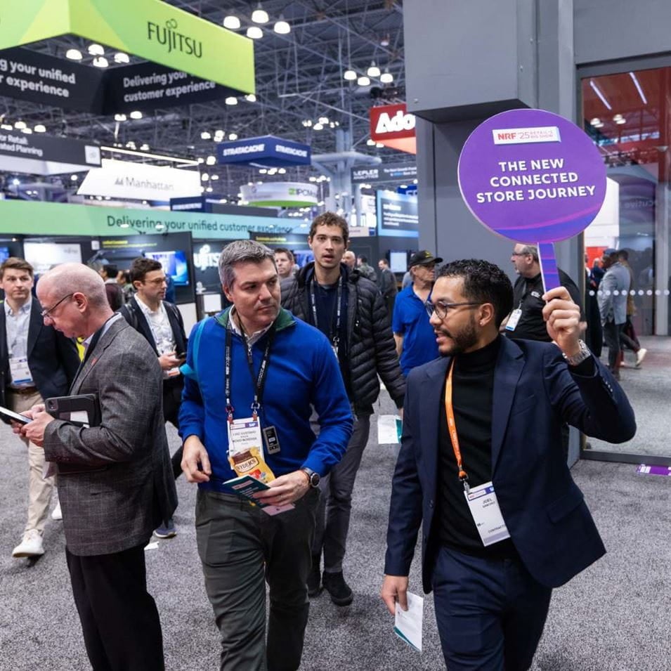 Guided tour at NRF 2025, a speaker holds a sign "The New Connected Store Journey" leading a group through the exhibition area.