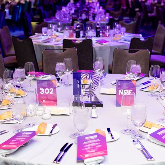 Table élégamment dressée pour la cérémonie des NRF Honors, avec menus, trophée en forme de "R", numéros de table et éléments de branding aux couleurs violet et rose.