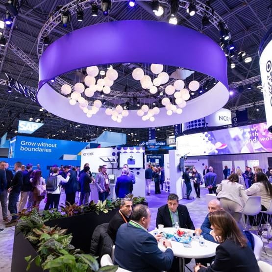 View of NRF 2025 show floor with visitors gathered around SAP and KPCYNYX booths under a large suspended light structure.